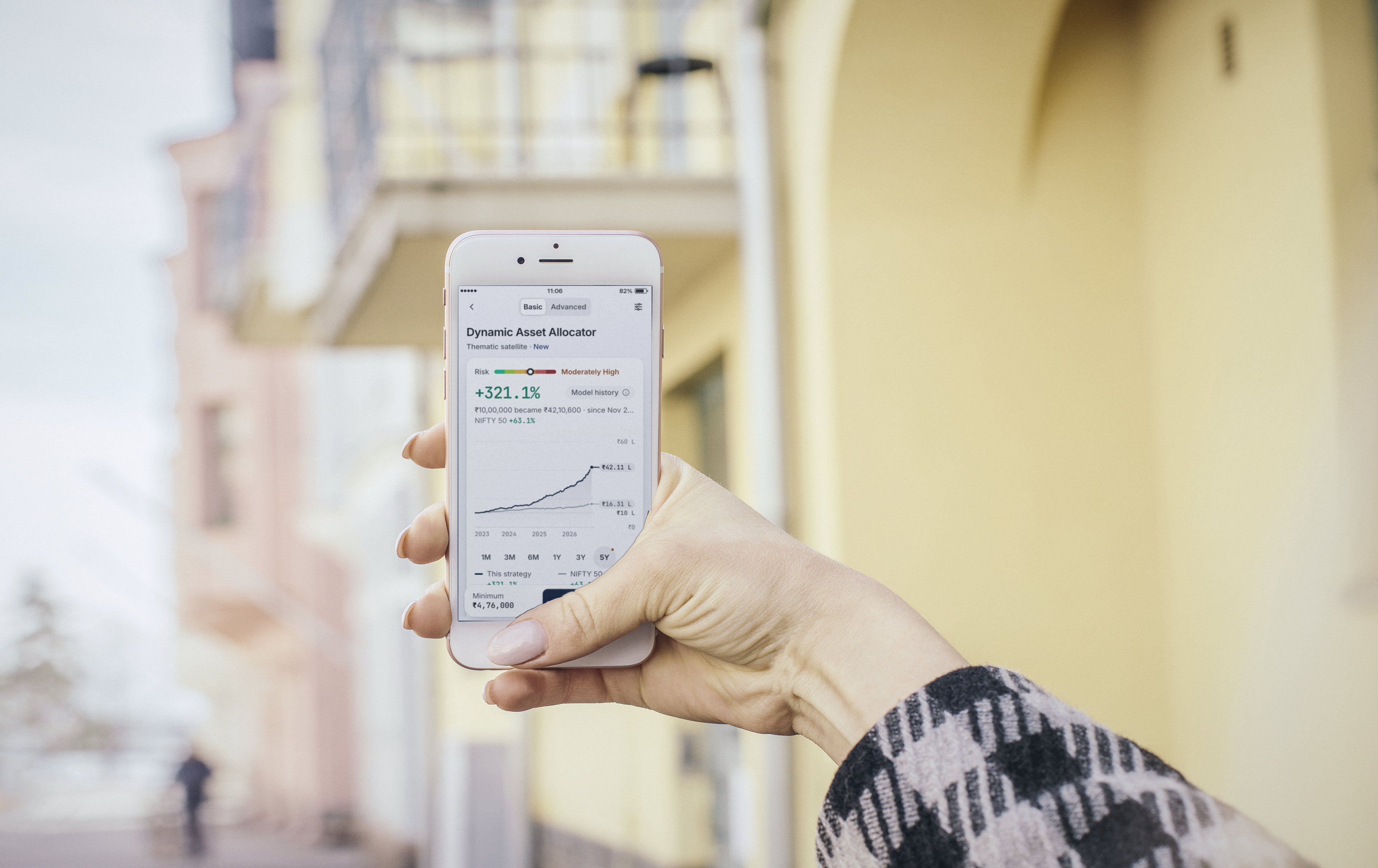 A hand holding a phone up in daylight on a city street, showing the Dynamic Asset Allocator strategy page with its model history chart